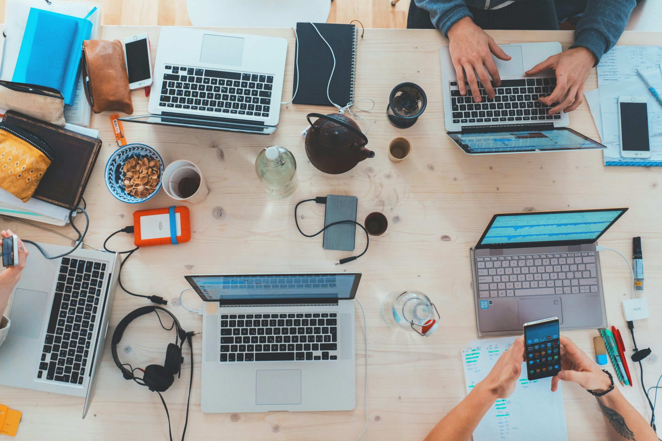 A bird's eye view of a busy desk, with multiple laptops, charging banks, a tea pot, drinks, and people working.